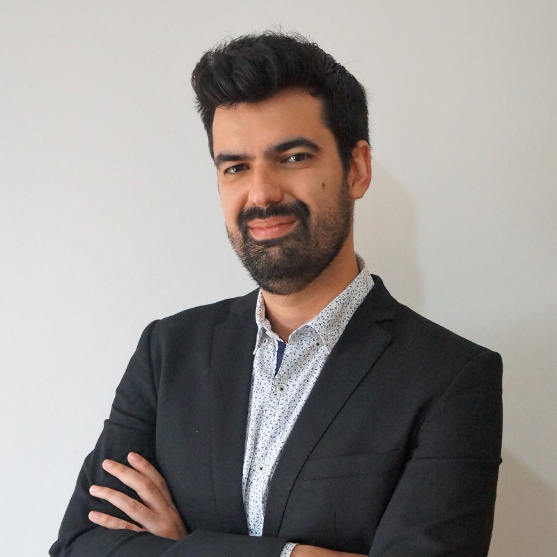 A professional portrait of a smiling man with a beard, wearing a black blazer and an AI-themed patterned shirt. He is standing indoors with a plain, light-colored background.