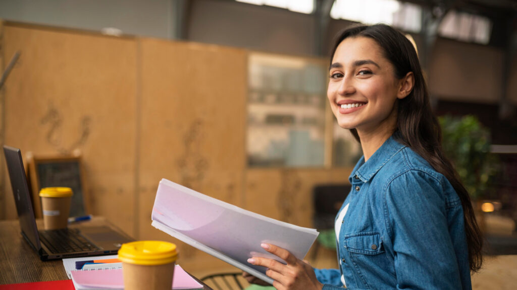 A smiling woman in a denim jacket holds papers outlining main competencies and sits at a desk with a laptop and coffee cups in an office with warm lighting and employability skills.