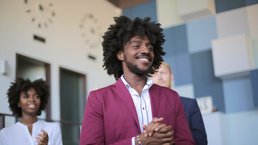 A confident young black man in a maroon blazer and white shirt, smiling and standing in a modern office, with his unique competencies apparent, and two colleagues slightly blurred in the background.