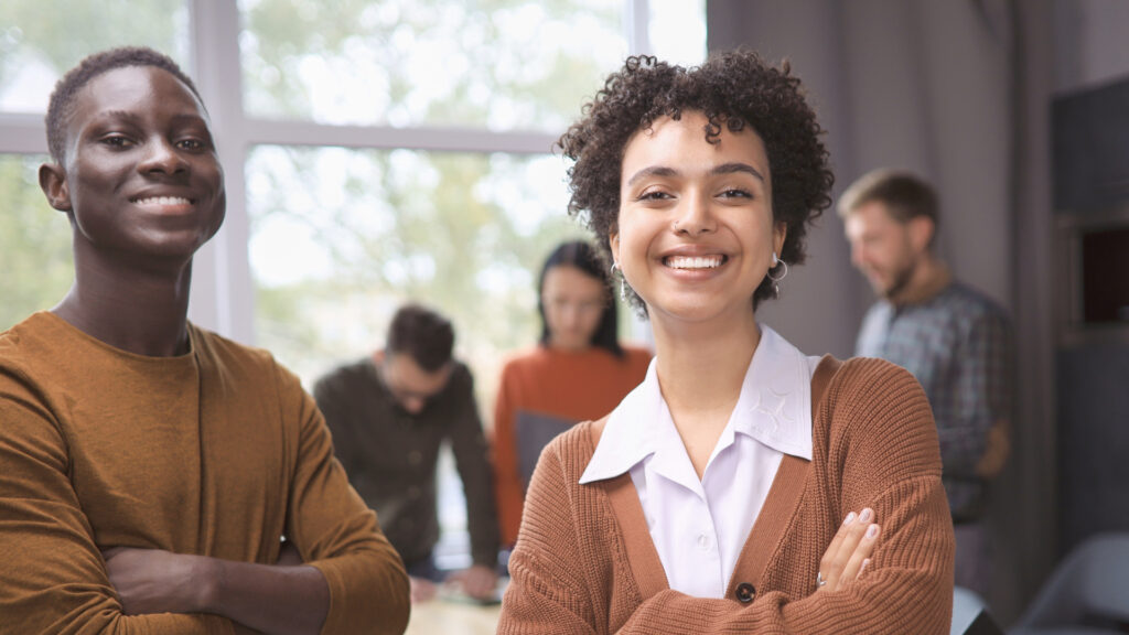 A young woman and a young man smiling at the camera, with three youth professionals discussing in the background in a bright, modern office.