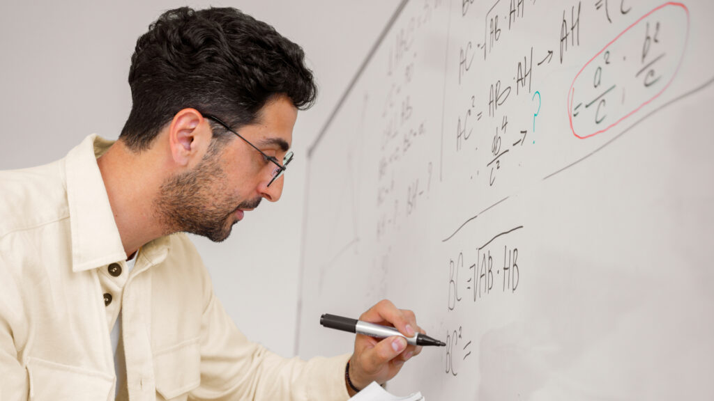 A young male teaching PISA in Brazil with curly hair, wearing glasses and a beige shirt, writes mathematical equations on a whiteboard using a black marker.